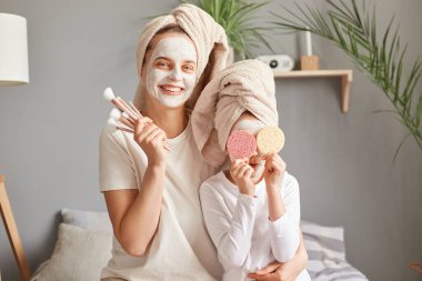 Olumlu gülümseyen anne ve yüz maskeli küçük kızı sabahları ellerinde toz fırçasıyla eğleniyorlar çocuk gözlerini süngerle kapıyor..