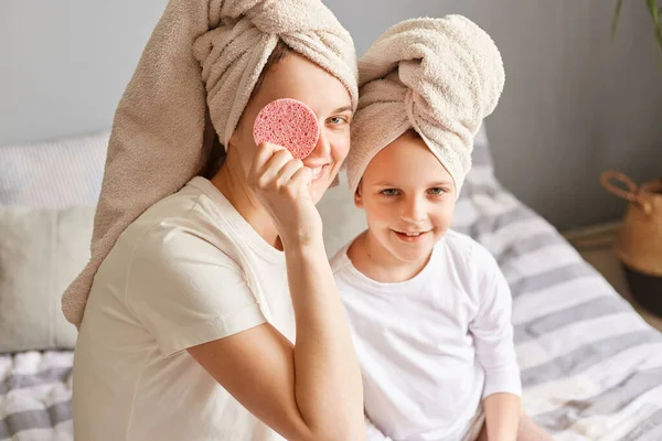 快乐的孩子 妈妈早上洗完澡后穿着毛巾坐在床上 让快乐的女人用化妆品海绵盖住眼睛 做美容手术 — 图库照片