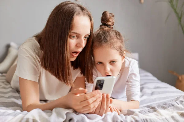 震惊的母亲和女儿使用手机 躺在床上 看着智能手机屏幕 大眼睛在网上购物 价格低廉 上网浏览 — 图库照片