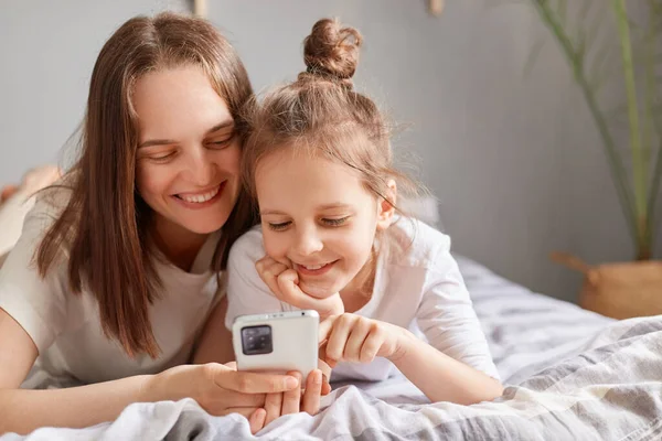 用手机微笑着妈妈和可爱的女儿 在床上放松 带着好奇的小女孩和妈妈一起看智能手机屏幕 浏览应用程序 — 图库照片