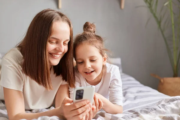 快乐的妈妈和可爱的女儿用手机 躺在床上 和妈妈一起看手机显示屏 上网购物 — 图库照片