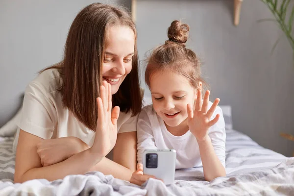 快乐快乐快乐的妈妈和女儿用手机 在床上放松地看着家里的智能手机屏幕上有视频通话 挥手打招呼 — 图库照片