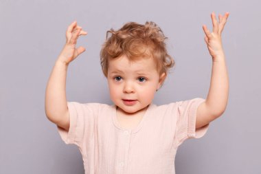 Sarışın, dalgalı tişörtlü, kollarını kaldırmış, gri arka planda ne kadar yalıtılmış poz vereceğini gösteren ciddi, küçük, beyaz bir kız çocuğu.