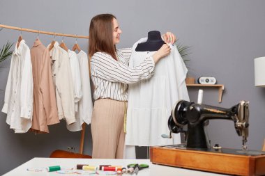 Kahverengi saçlı terzi kumaşa iğne takıyor atölyesinde çalışıyor, düzen içinde çalışıyor, şık kıyafetler dikiyor, iş yerinin yanında duruyor..