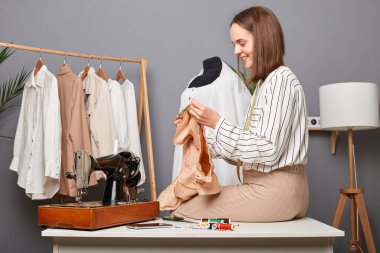 Dikiş atölyesindeki beyaz kadın portresi. Yeni elbisenin detaylarını tutmak, masaya oturmak, gülümsemek, tasarımcı yeni koleksiyon yaratmak..