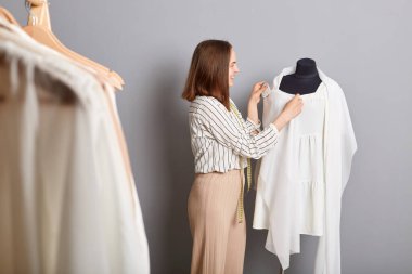 Yeni sezon için atölyede çalışan iğneler kullanılarak gri arka planda dikilen yeni elbiseler ve yeni elbiseler üzerinde duran konsantre gülümseyen kadın lağımı..