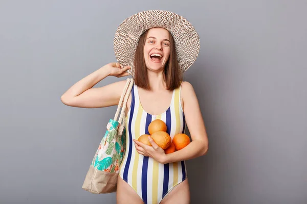 夏の旅行 笑顔笑って若いです大人の女性を身に着けていますストライプ水着とわら帽子保持新鮮な果物楽しみます休暇を取ります隔離された灰色の背景 — ストック写真