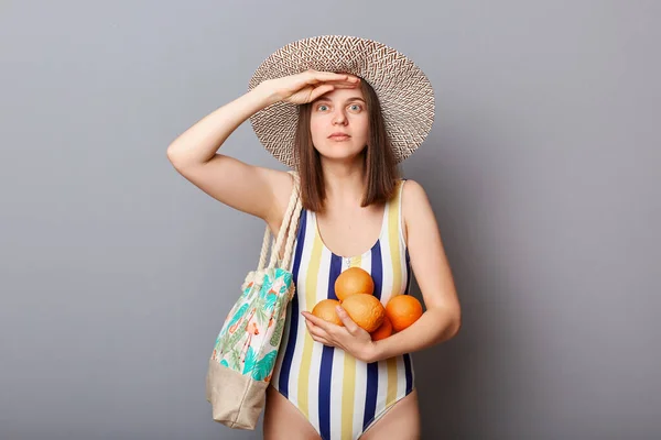驚くべき驚きの女性を身に着けていますストライプ水着とわら帽子隔離されたグレーの背景立ちで新鮮なオレンジと手を保ちます額の近くに見てはるかに — ストック写真