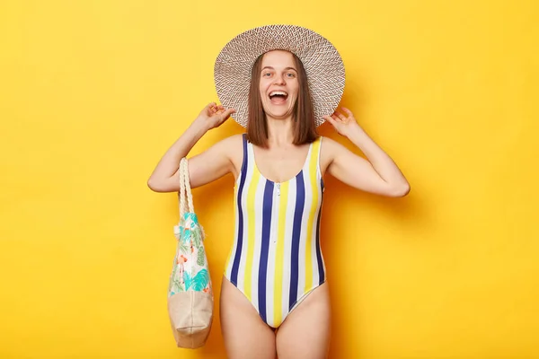 縞模様の水着と麦藁帽子を身に着けている素晴らしい女性隔離された黄色の背景保持バッグは笑い続けます楽しい休暇夏休み — ストック写真