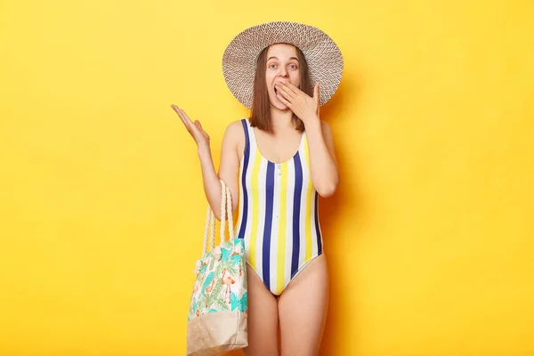 あなたの広告のための空のスペースを指している縞模様の水着とわら帽子隔離された黄色の背景を身に着けている素晴らしい驚きの女性 空白の壁を示す — ストック写真
