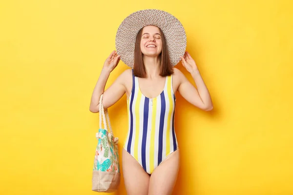 縞模様の水着と麦藁帽子を身に着けている楽しい笑顔の女性閉鎖的な目で立っている黄色の背景夏休みを楽しんで熱帯休暇 — ストック写真