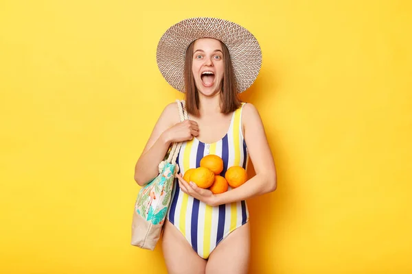 縞模様の水着と麦藁帽子を身に着けている非常に驚くべき女性は袋と新鮮なオレンジと立って黄色の背景を隔離幸せな休暇を楽しんで叫んで — ストック写真