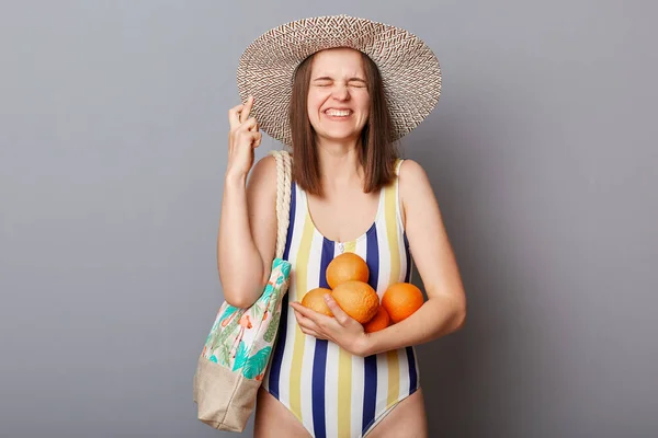 希望の笑顔幸せな女性身に着けているストライプカラフルな水着スタンディングでバッグとオレンジ隔離された上にグレーの背景交差指良い休暇を持ちたい — ストック写真