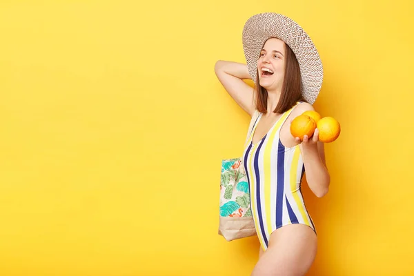 黄色の背景に立ってオレンジを持って幸せそうに見える彼女の休暇中においしくて健康的な軽食を楽しむことを期待しています — ストック写真