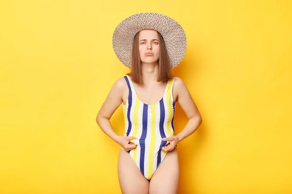 絶望女性オンリゾート身に着けていますで縞模様の水着と藁帽子ポージング孤立上の黄色の背景ショー彼女の脂肪腹必要性へ緩い重量ない準備彼女の体のための夏 — ストック写真