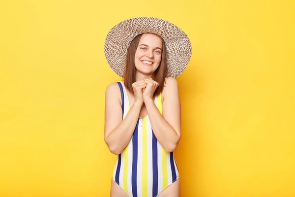 黄色の背景に隔離されながらも 縞模様の水着と太陽の帽子を身に着けて明るい表情の女性は カメラを直接見て ビーチで平和な一日を楽しんで — ストック写真