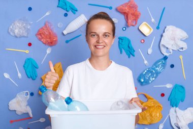 Kirlilik çevreye zarar verir. Mavi duvarda izole edilmiş beyaz gömlekli, gülen bir kadın çöp tenekesi ve çöp tenekesiyle etrafı temizliyor..