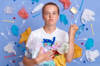 Gönüllüler çöpleri temizler. Atıkları azaltmak için malzemeleri yeniden kullanın. Mavi duvarda izole edilmiş beyaz gömlek giyen kızgın bir kadın. Etrafı çöp dolu, yumruğunu kaldırmış..