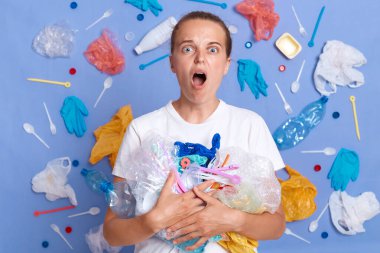 Şok olmuş beyaz gömlekli bir kadın mavi duvarda izole edilmiş, etrafı çöplerle çevrili büyük gözlerle kameraya bakıyor ve ağzı açık bir şekilde plastiğin gezegenimize zarar verdiğini duyuyor..