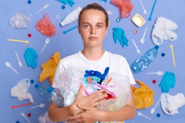 Etrafı temizliyorum. Kirlilik dünyayı etkiler. Çöp çevreye zarar verir. Katı, otoriter, beyaz tişört giyen, etrafı çöplerle dolu, mavi duvarda izole bir kadın..