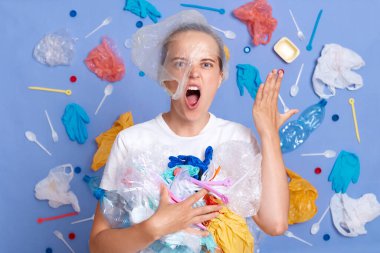 Kızgın, agresif, beyaz tişörtlü bir kadın mavi duvarda izole edilmiş, etrafı çöplerle çevrili nefretle bağıran çılgın yüzlü, gezegen kirliliğinden dolayı umutsuzluğa düşmüş bir kadın..