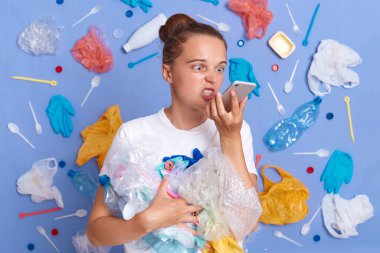 Beyaz gömlekli çılgın kadın mavi duvarda izole edilmiş. Etrafı çöp dolu. Sesli mesaj kaydediyor..