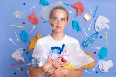 Eko için tekrar kullan. Temizlik için gönüllü ol. Gezegenimizi koru. Mutsuz bir kadın, mavi duvarda izole edilmiş beyaz tişört giyiyor. Etrafı çöplerle çevrili ve ciddi bir yüzle kameraya bakıyor.