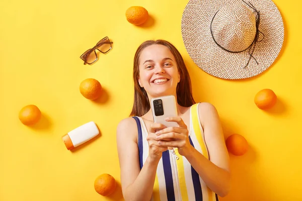 stock image Smiling woman wearing striped swimming suit lying with oranges sunscreen on yellow background using mobile phone with happy expression browsing internet.