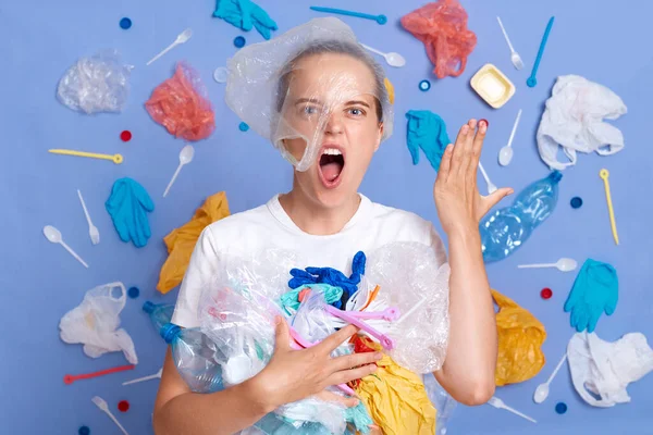 Kızgın, agresif, beyaz tişörtlü bir kadın mavi duvarda izole edilmiş, etrafı çöplerle çevrili nefretle bağıran çılgın yüzlü, gezegen kirliliğinden dolayı umutsuzluğa düşmüş bir kadın..