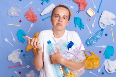 Belirsiz bir kadın mavi duvarda izole edilmiş beyaz gömlek giyiyor. Çöp yığını ve akıllı telefonuyla elinde kameranın omuz silkmesini seyrediyor..