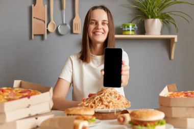 Lezzetli yemek. Çevrimiçi sipariş, hızlı teslimat. Beyaz tişörtlü, gülümseyen bir kadın mutfakta abur cuburla oturmuş akıllı telefonu boş ekranlı gösteriyor, reklam için kopyalama alanı var..
