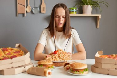 Kafası karışmış, beyaz tişörtlü bir kadın mutfakta fast food yiyor kafası karışmış suratlı sağlıksız tabaklara bakıyor, çok fazla yemek sipariş ediyor..
