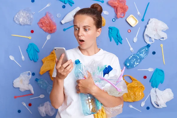 Reduce garbage pollution. Cleaning up planet. Shocked surprised woman wearing white shirt isolated on blue wall with garbage around looking at cell phone screen with open mouth.