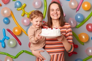 Çizgili elbiseli, gri duvara yaslanmış gülümseyen bir kadın balon süslemeli, elinde küçük kızı ve doğum günü pastası, lezzetli bir tatlı..