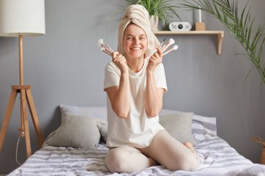 Kafasında havluyla son derece mutlu bir kadın evinde yatağında yeni kozmetik fırçalar tutuyor gülümseyerek kameraya bakıyor..