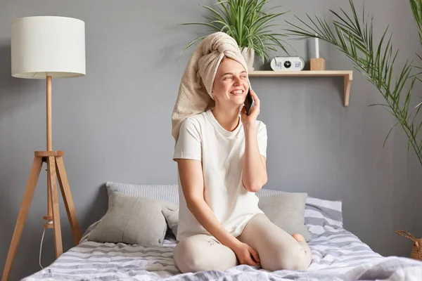 ベッドに座っている間に携帯電話を使ってシャワータオルに包まれ 国内服を着て楽しい会話をしている若い女性 — ストック写真