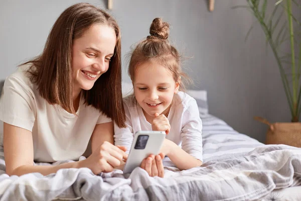 用电话微笑着妈妈和可爱的女儿 在床上放松一下 带着快乐的表情看着智能手机屏幕的小女孩 — 图库照片