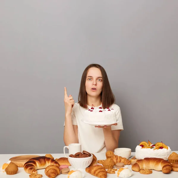 Inspired woman wearing white T-shirt isolated over gray background having cheat meal holding cake raised her finger having idea pointing at advertisement area.