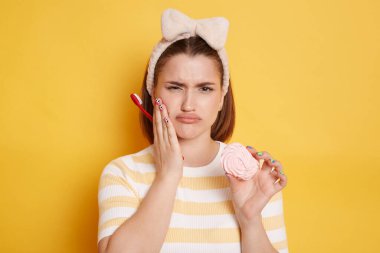 Sağlıksız bir kadın dişleri için zararlı şekerleme yiyor ve acı çekiyor, diş fırçası tutuyor, ağzına dikkat etmeli, sarı arka planda izole edilmiş günlük çizgili tişört giymeli..