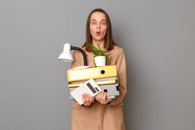 Şaşırmış bir kadın, elinde karton kutuyla işini kaybetmiş gri arka planda izole edilmiş bir şekilde kameraya bakarak ağzı açık bir şekilde poz veriyor..