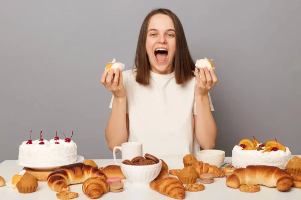 stock image Extremely happy cheerful woman wearing white T-shirt isolated over gray background sitting at festive table among desserts holding cakes screaming with excitement.