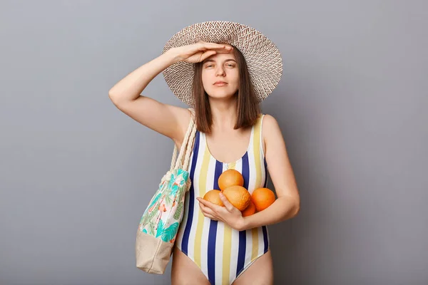 真剣な女性身に着けています水着絶縁上の灰色の背景立っていますとともにオレンジキープ手近く額見ますはるかに待っています彼女のトロピカル休暇 — ストック写真