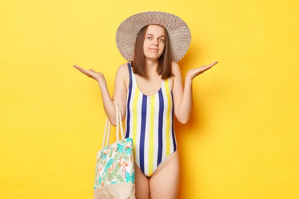 混乱した表情で立って黄色の背景に隔離された水着を身に着けている謎の不確定な女性肩は休暇の出発に関する情報を知らない — ストック写真