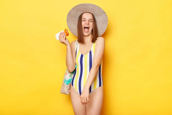 彼女の腕の上に日焼け止めを適用し 黄色の背景に隔離された水着を身に着けている非常に幸せな女性叫んで幸せ日光浴を楽しむ — ストック写真