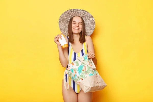 満足のいく女性が水着を着て黄色の背景に立ってビーチバッグでわら帽子彼女の保護Spfクリームを保持している彼女の品質を楽しんで — ストック写真