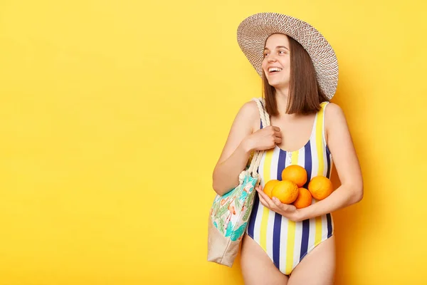 明るい女性が水着姿で黄色の背景に孤立し 新鮮なオレンジが広告エリアを離れて見て肯定的な感情で微笑む — ストック写真