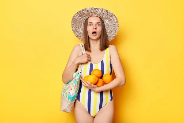 黄色の背景に隔離された水着姿の衝撃的な女性保持オレンジあります驚いています高い価格のために果物見カメラでオープン口 — ストック写真