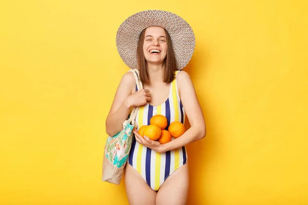黄色の背景に隔離された水着を身に着けている幸せな喜びの女性は喜んで彼女の休暇を楽しんで笑って 新鮮なオレンジはトロピカルフルーツが好き — ストック写真
