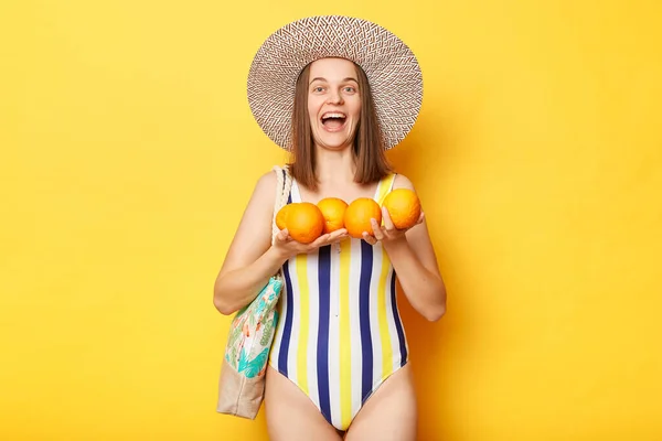 黄色の背景に隔離された縞模様の水着を身に着けている興奮した女性提供新鮮なオレンジ叫んで満足している素晴らしい休暇 — ストック写真