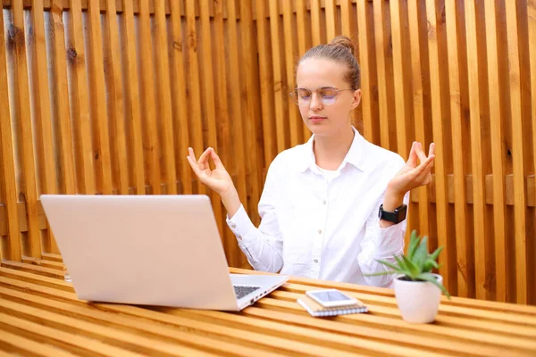 Aşırı çalışan, beyaz gömlekli rahat bir kadın dışarıda bir kafede oturmuş laptopla çalışıyor sakinleşmeye çalışıyor, yoga yapıyor, nefes alıyor..